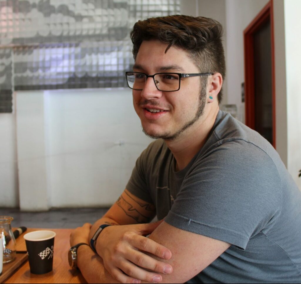 Matt Glaman seated at a table with coffee, smiling and looking off frame